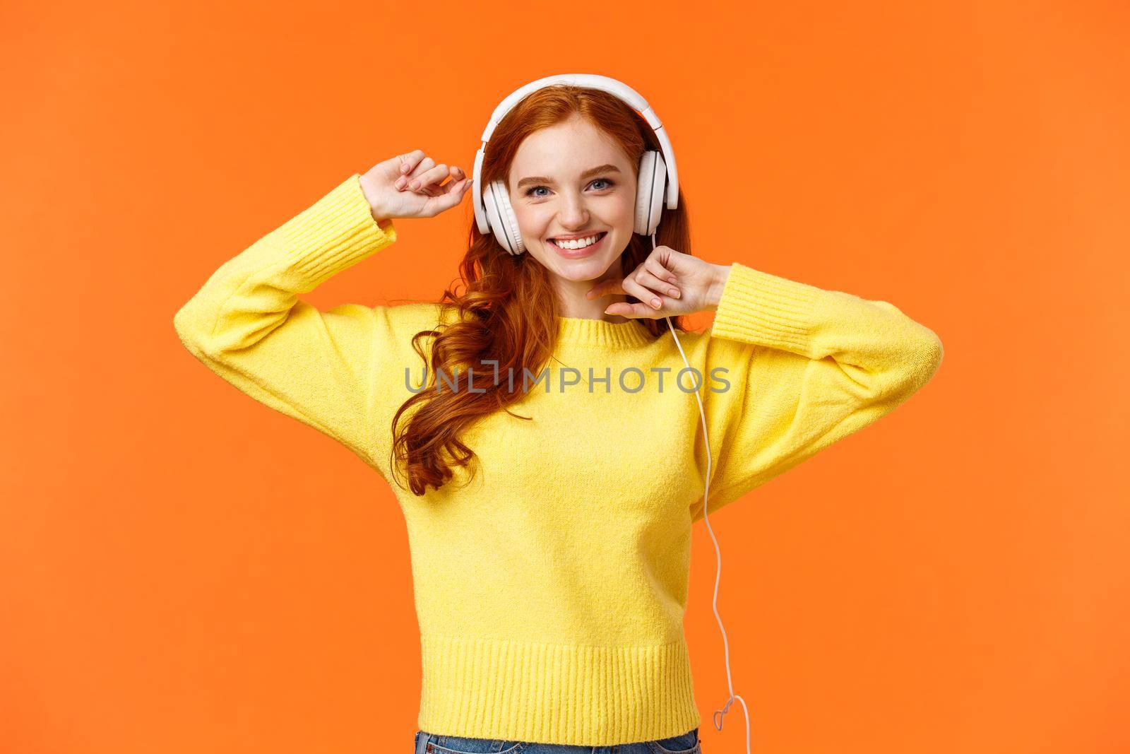 Happy and carefree excited redhead hipster girl like her new headphones, dancing with hands lifted up and smiling enjoy listening music over orange background, boost mood with favorite song.