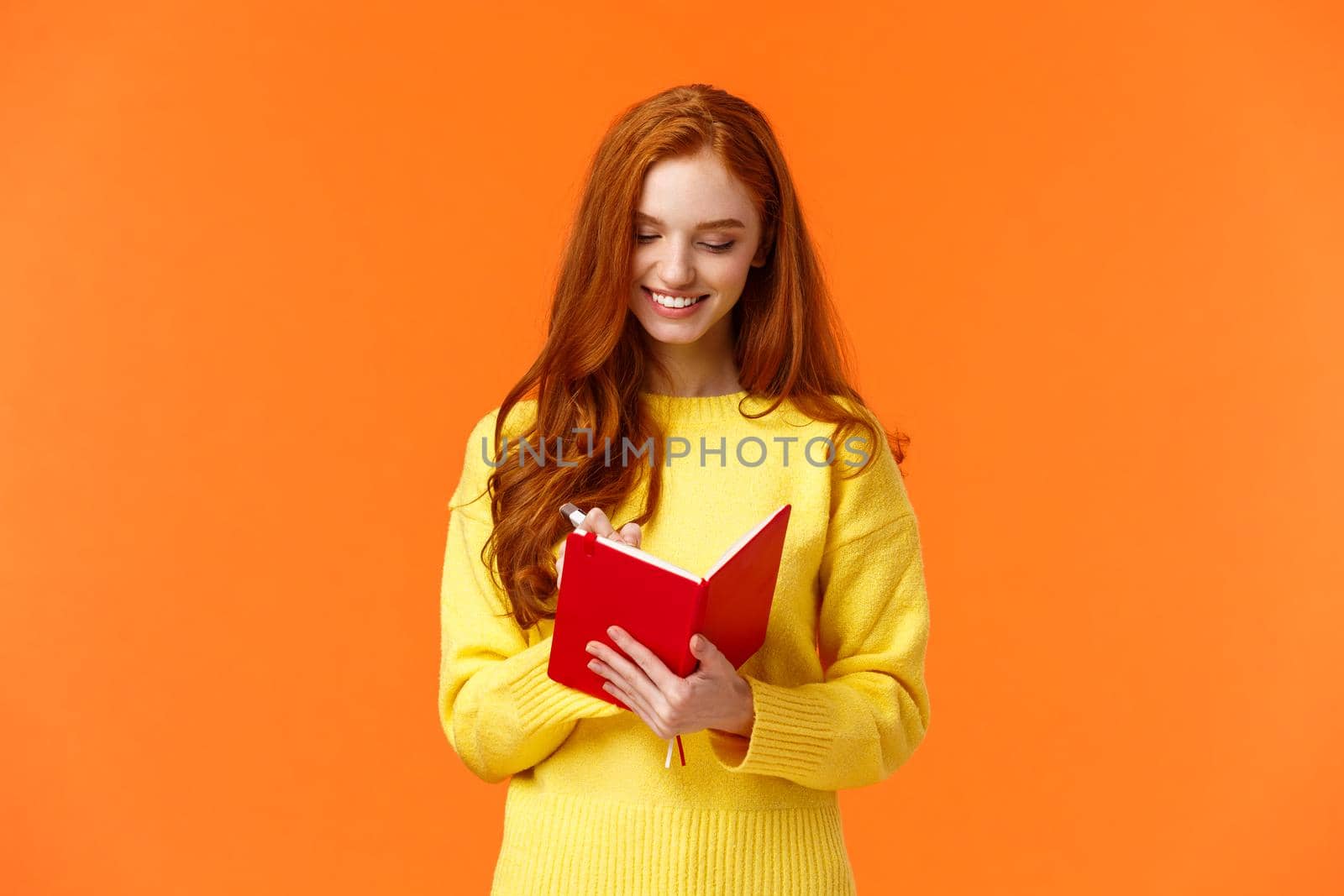 Waist-up portrait attractive redhead girl 20s write in her diary, fill-in to-do list or personal schedule in new notebook, smiling as having everything planned, standing orange background carefree by Benzoix