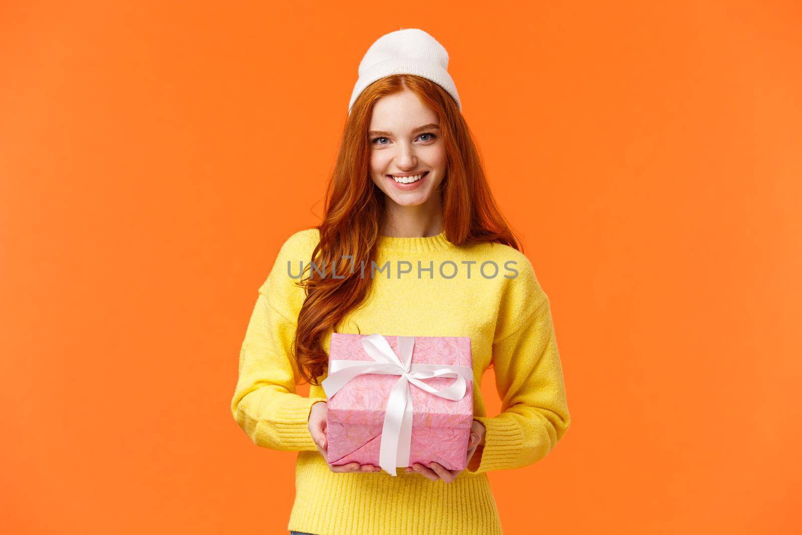 Cute redhead european woman prepare gift, wrapped present and giving it on christmas holidays, wearing winter beanie, smiling joyfully as congratulating someone, orange background.