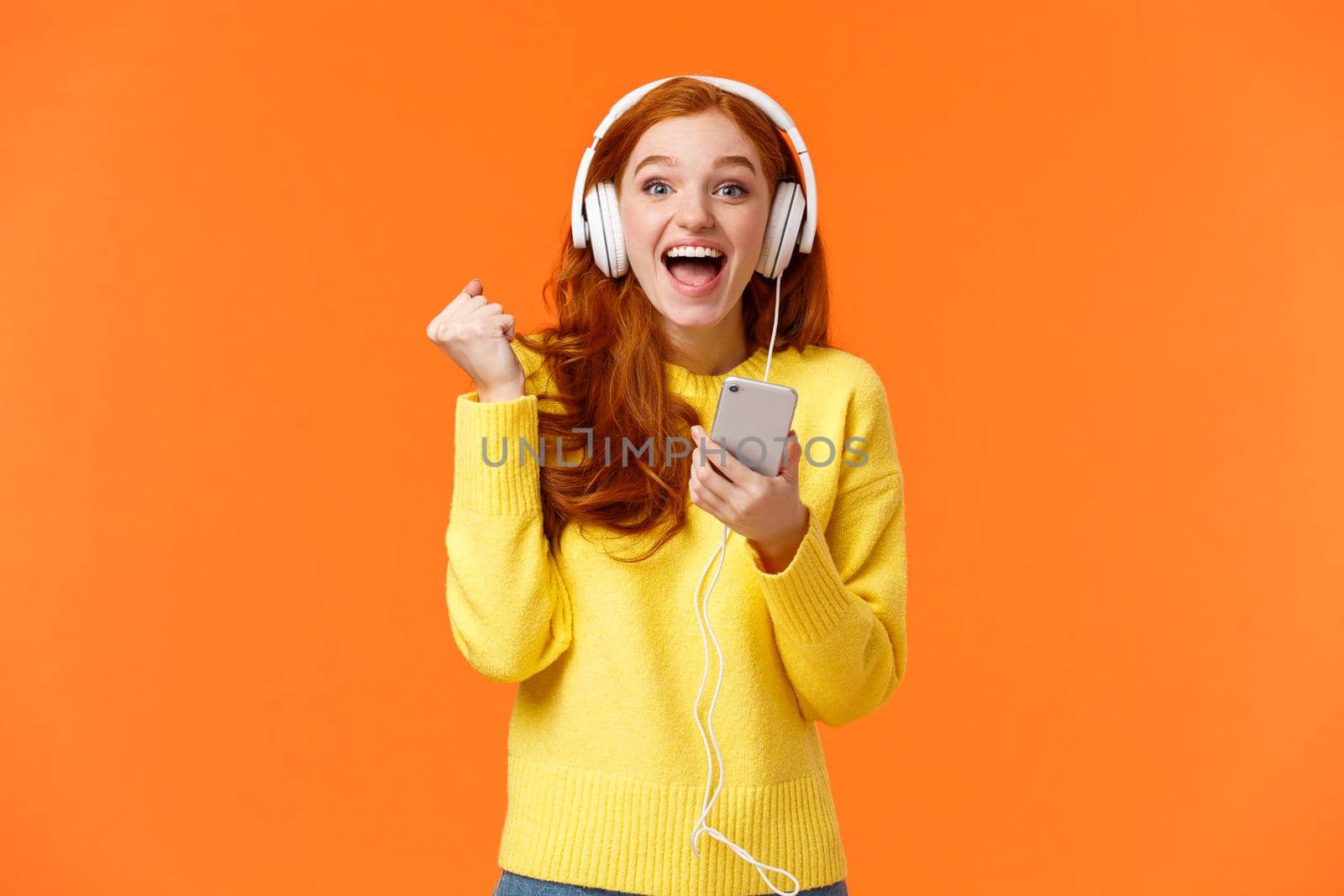 Hooray yes new song. Attractive cheerful and excited redhead woman fist pump in joy and positive emotions, wearing headphones, holding smartphone, smiling camera happily, orange background.