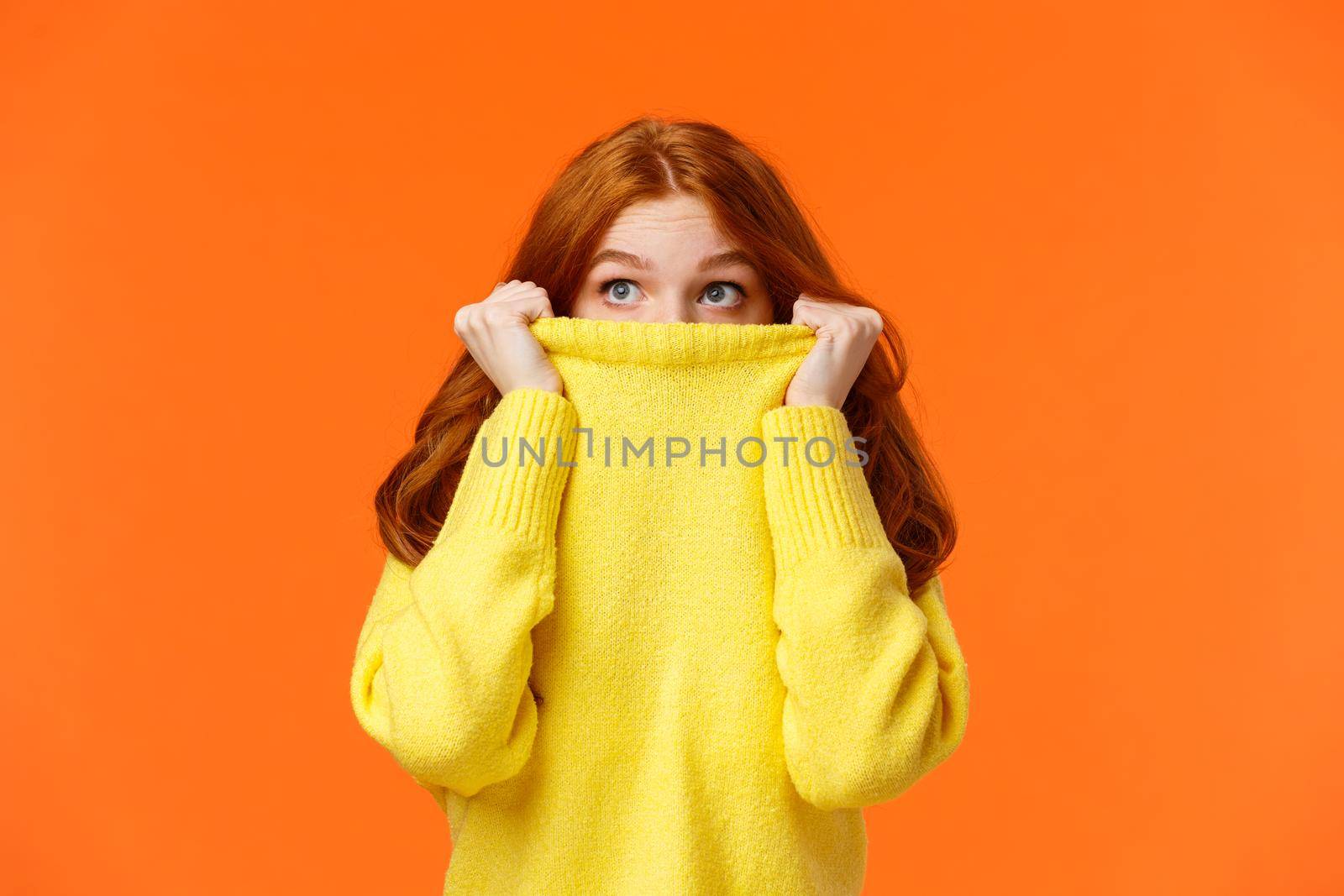 Scared cute teenage redhead girl pulling sweater on face and peeking at upper left corner with frightened eyes, glancing sideways, watching horror movie alone, orange background.