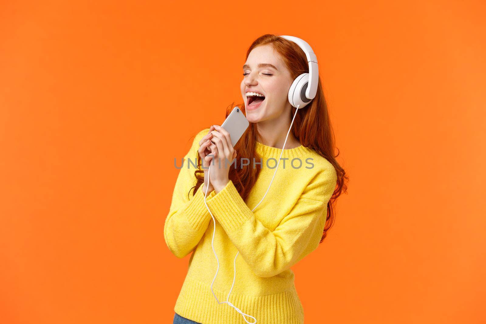Girl carried away while listen favorite track. Pretty redhead female enjoy music in new headphones parents bought for birthday, holding smartphone close eyes and singing in microphone as if karaoke.
