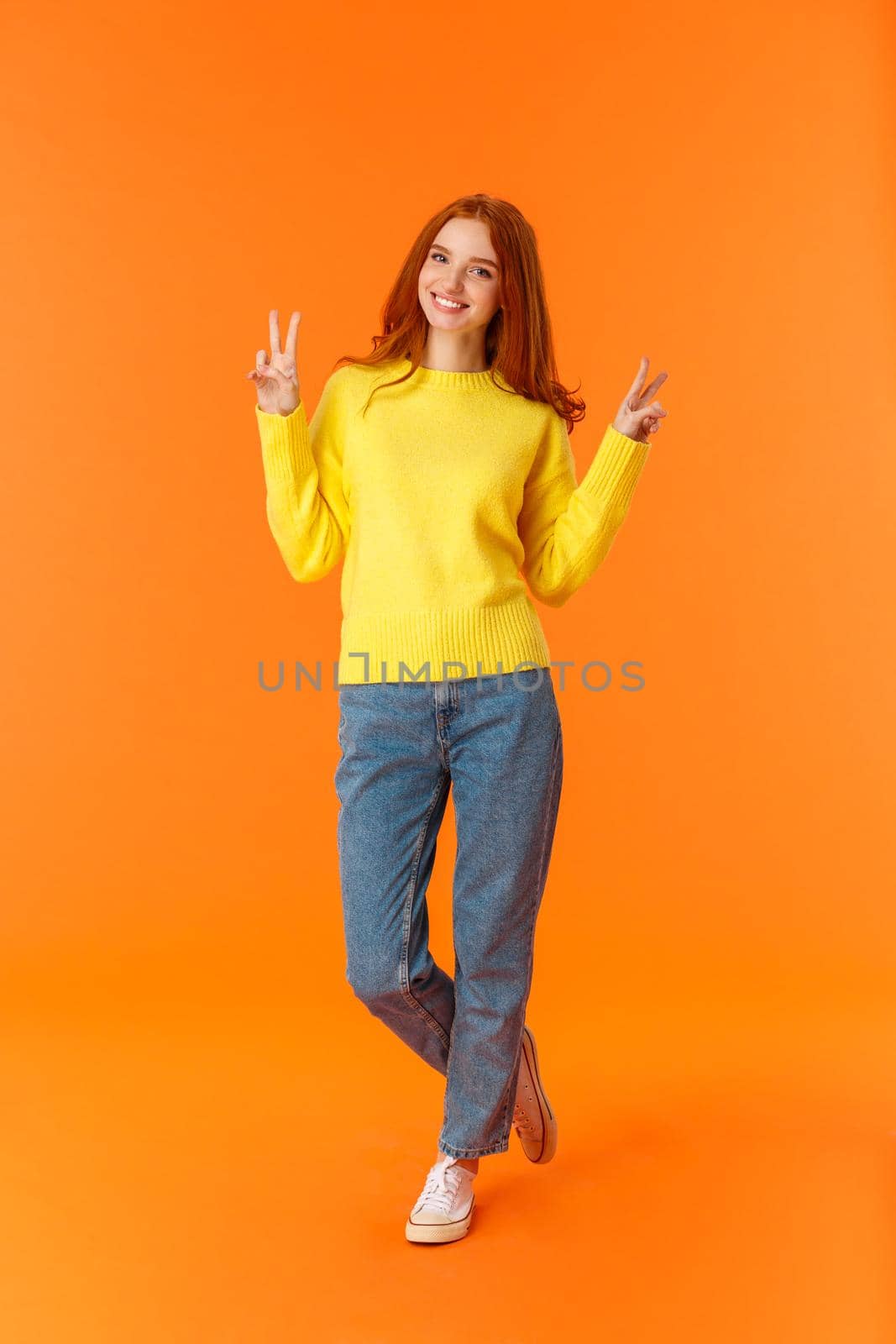 Vertical full-length cute lovely redhead teenage girl in jeans and winter yellow sweater standing with peace signs over orange background, smiling posing express carefree joy emotions by Benzoix