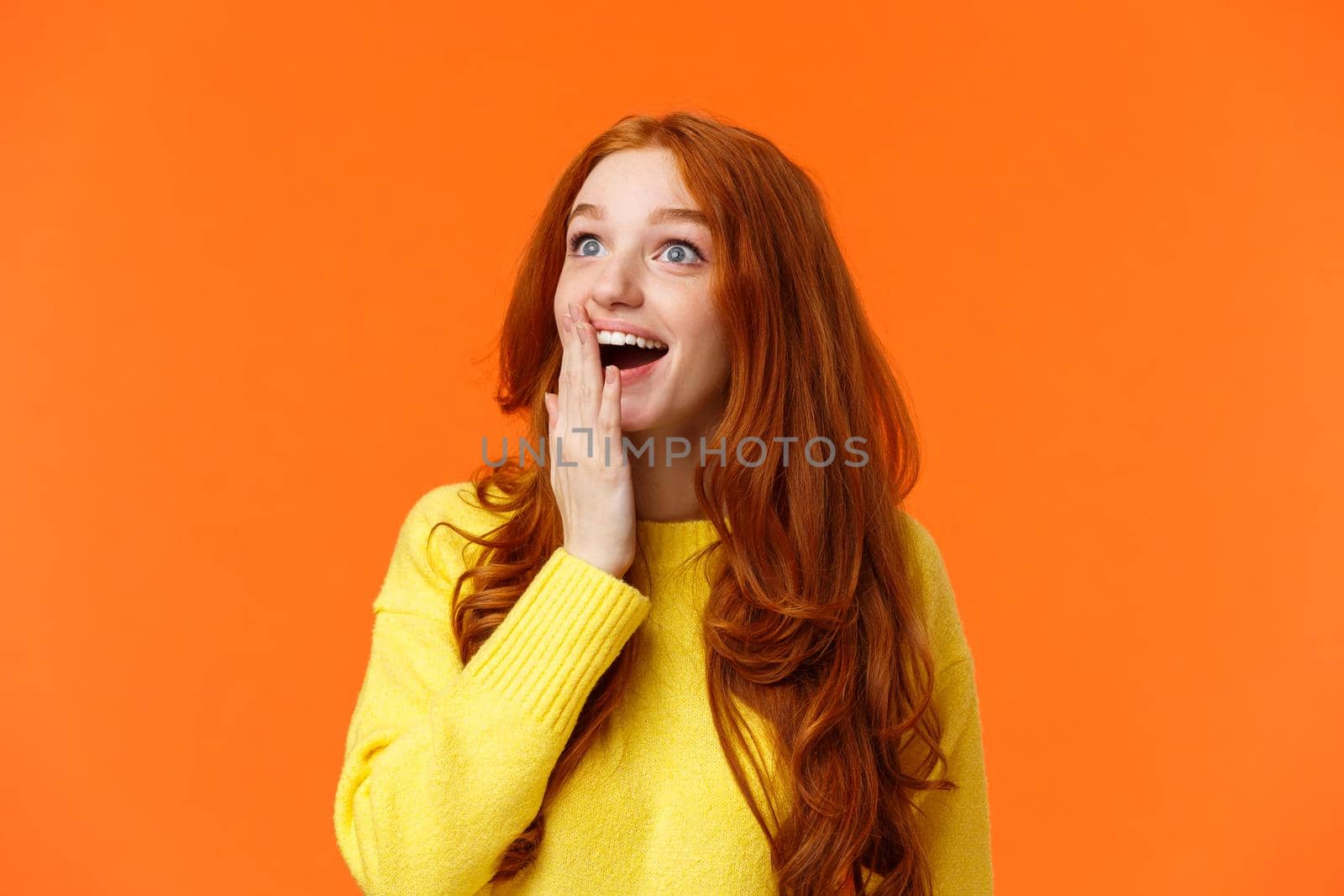 Fascinated and amiring cute redhead woman looking impressed and excited at upper left corner, smiling see breathtaking view, cover opened mouth palm thrilled, losing speech happy, orange background.