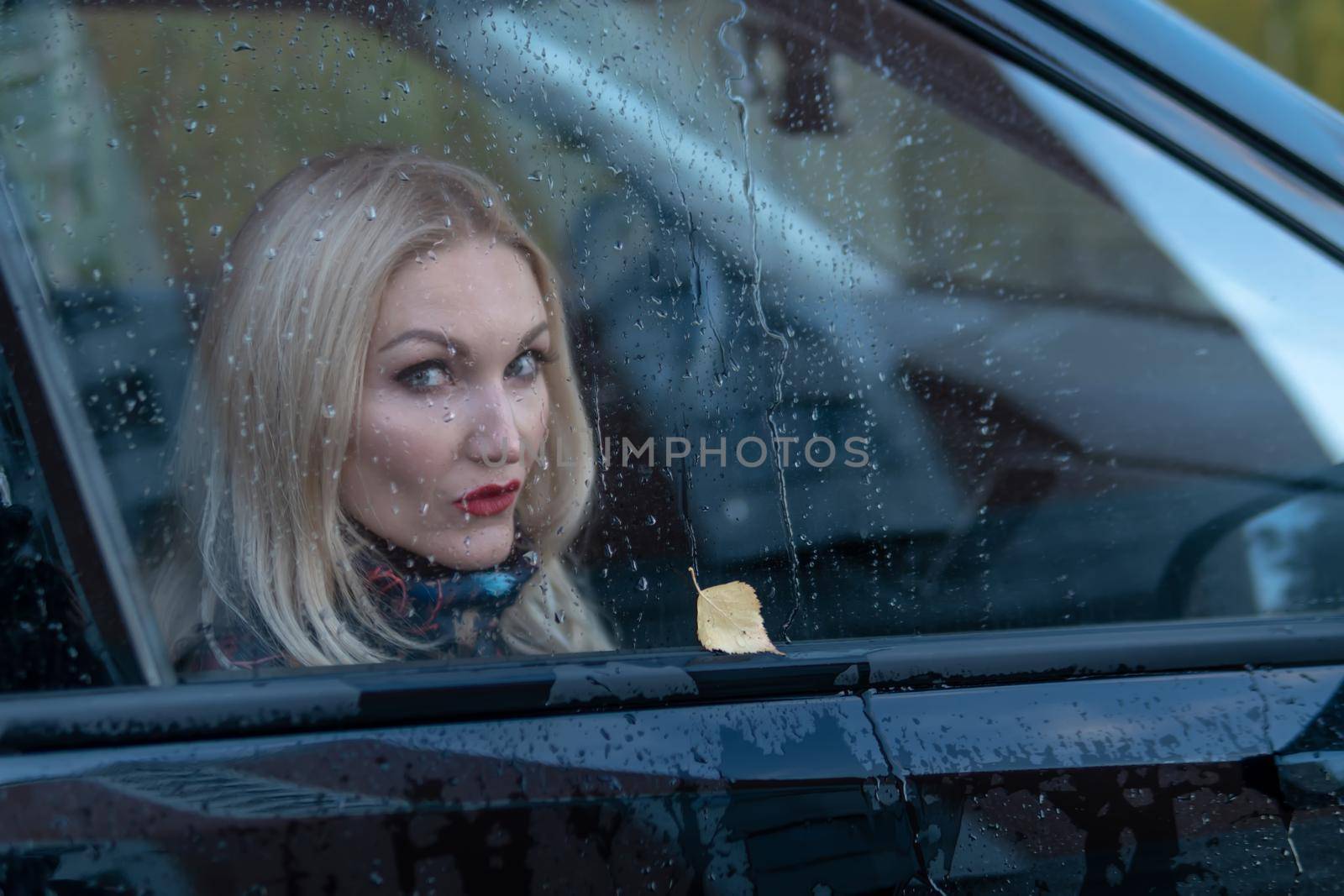 watching through the rainy glass, a girl drops from the car window on the rain, with a beautiful appearance in a black car. looking at camera by 89167702191