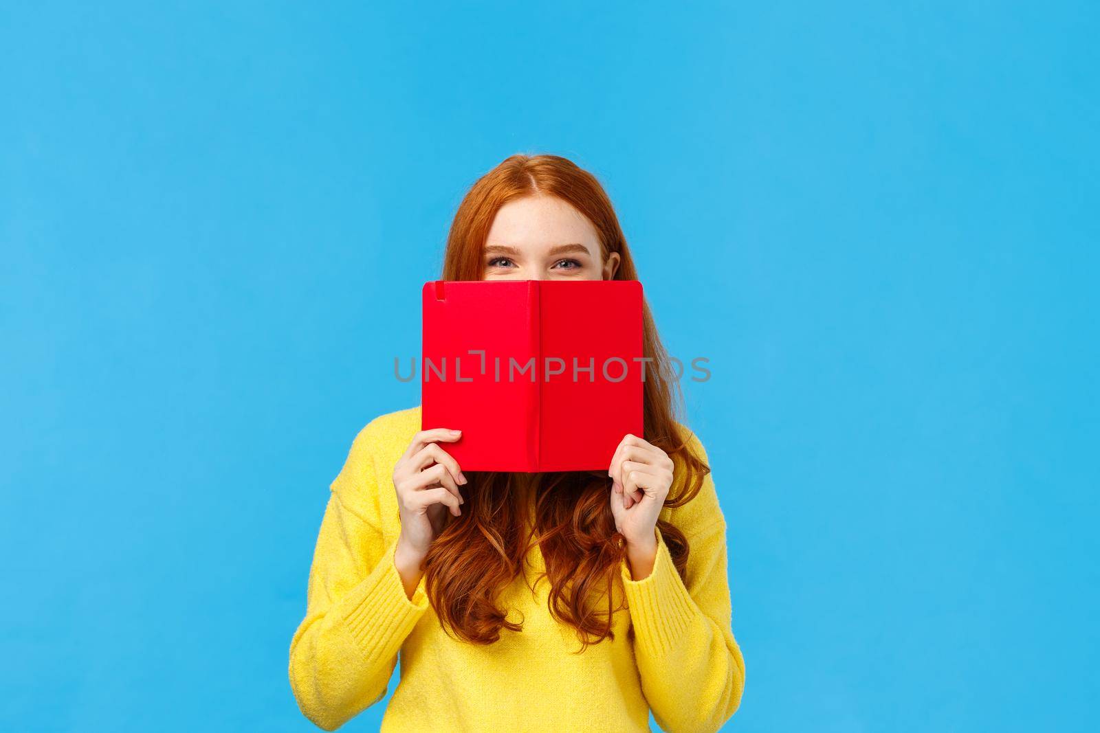 Sassy and creative happy charismatic redhead woman, teenager hiding face behind red notebook and smiling, grinning sly, hide from friend what she wrote in diary, standing blue background by Benzoix