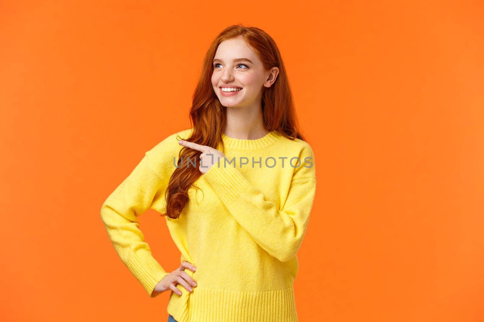 Dreamy, energized good-looking redhead female in yellow sweater, found excellent holiday gift, consumer pointing left, looking satisfied, smiling as picking product, make decision, orange background.