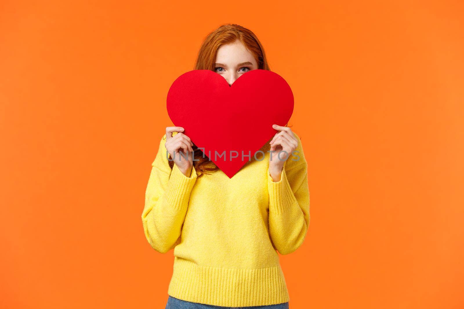 Cute romantic and blushing girl express her feelings on valentines day, hiding face shy behind big heart sign and peeking at camera, giving lovely gift, standing orange background tender by Benzoix