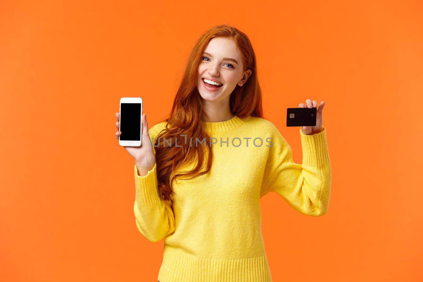 Shopping, online and finance concept. Cheerful young redhead female opened bank account, holding smartphone and credit card, smiling, recommend use online shopping application.
