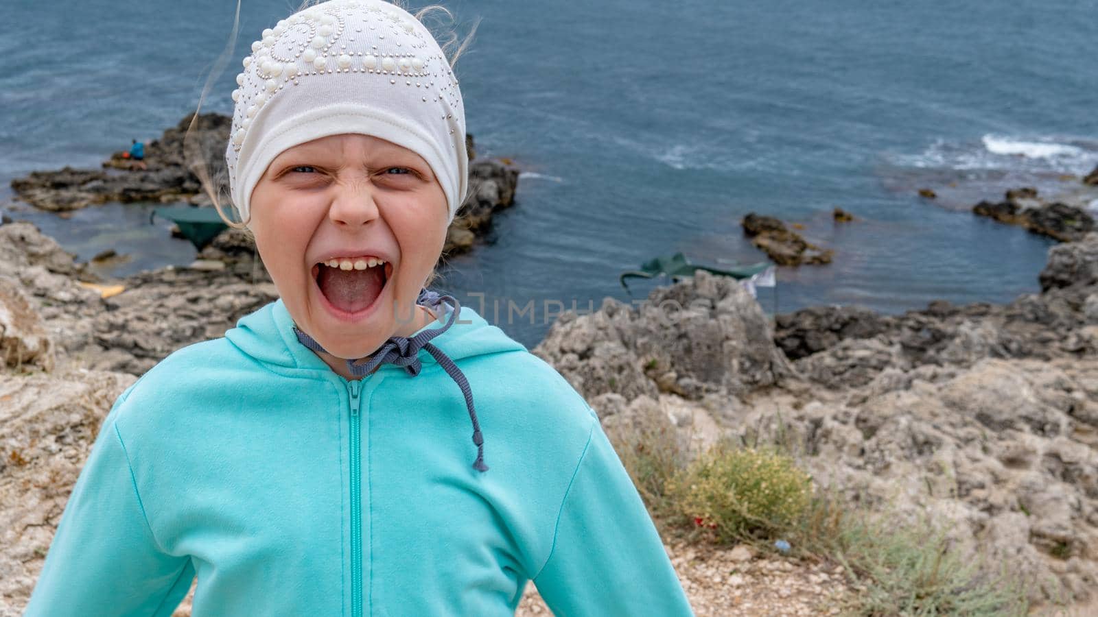 Girl at sea screams, rejoices in a jacket around the mountain, a girl in a hat on the beach in the summer in a jacket