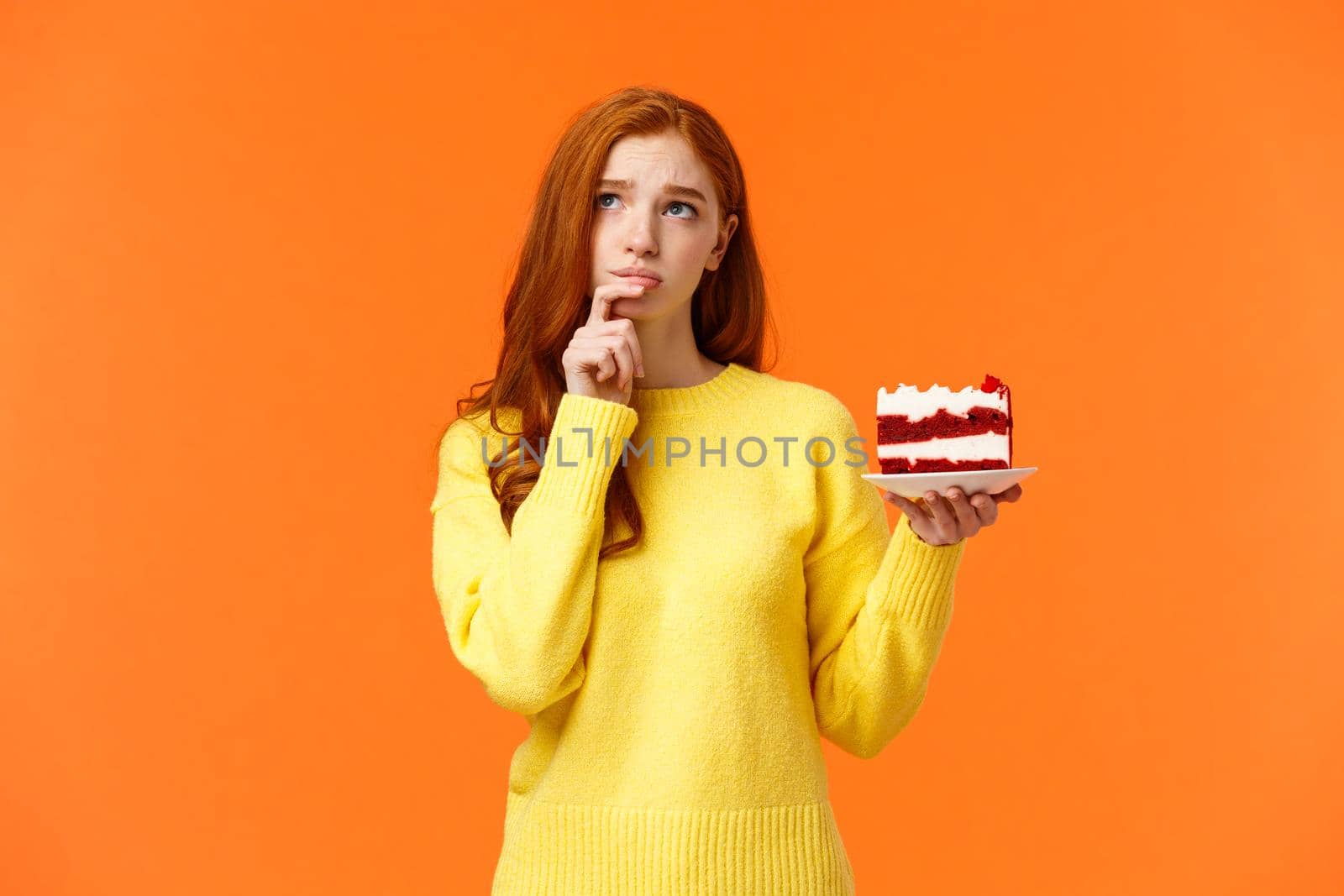 Worried, concerned silly young redhead girl taking care her body, calculating calories, being diet and want eat delicious cake, holding piece of dessert, look up thoughtful and troubled, orange wall.