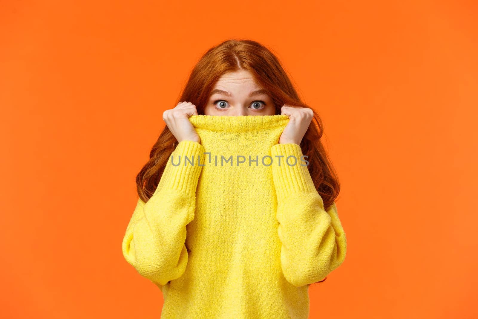 Surprised and astonished cute curious redhead female pulling sweater collar on face and popping eyes at camera with amazed expression, found something interesting, orange background by Benzoix