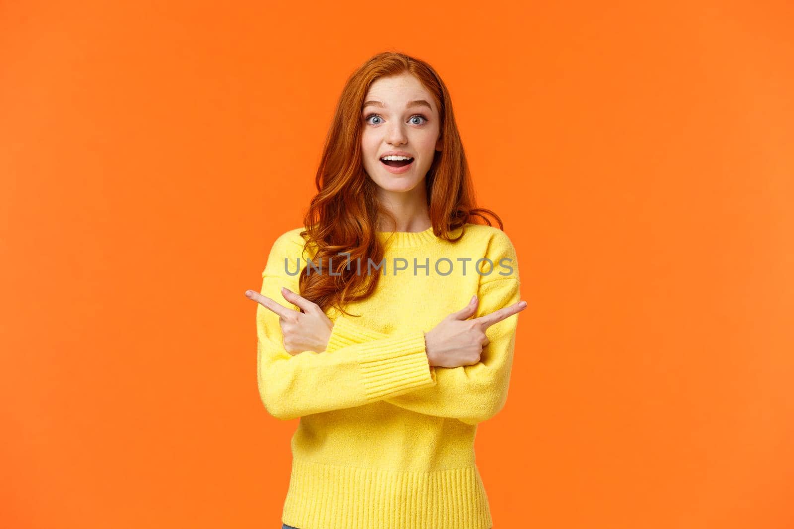 Amused dreamy and cute redhead girl asking advice in shopping mall, buying holiday presents, want valentines day gift, pointing sideways left and right, smiling upbeat, orange background.
