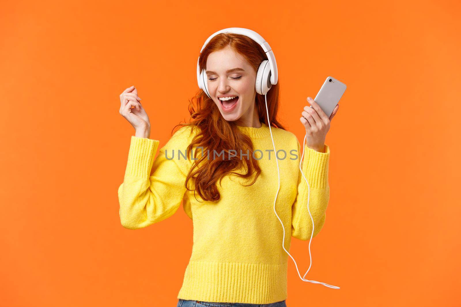 Technology, emotions and gadgets concept. Cheerful good-looking redhead woman dancing with hands up, closed eyes, smiling joyfully, enjoy awesome music sound in headphones, hold smartphone by Benzoix