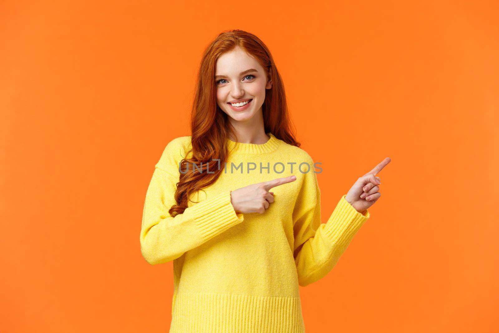 Time to prepare for holidays. Cheeky gorgeous smiling girl with red curly hair, pointing right, advice what shop visit, recommend online store for winter goods, standing orange background.