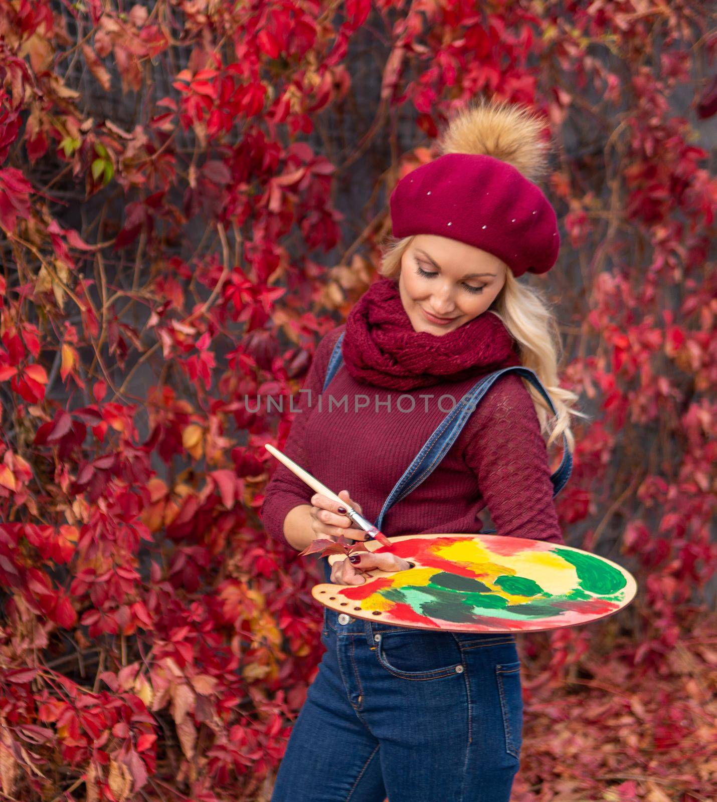 The artist paints red maple leaves on the leaves with red paints in blue jeans and a burgundy sweater. Cheerful looking at camera enjoy concept by 89167702191