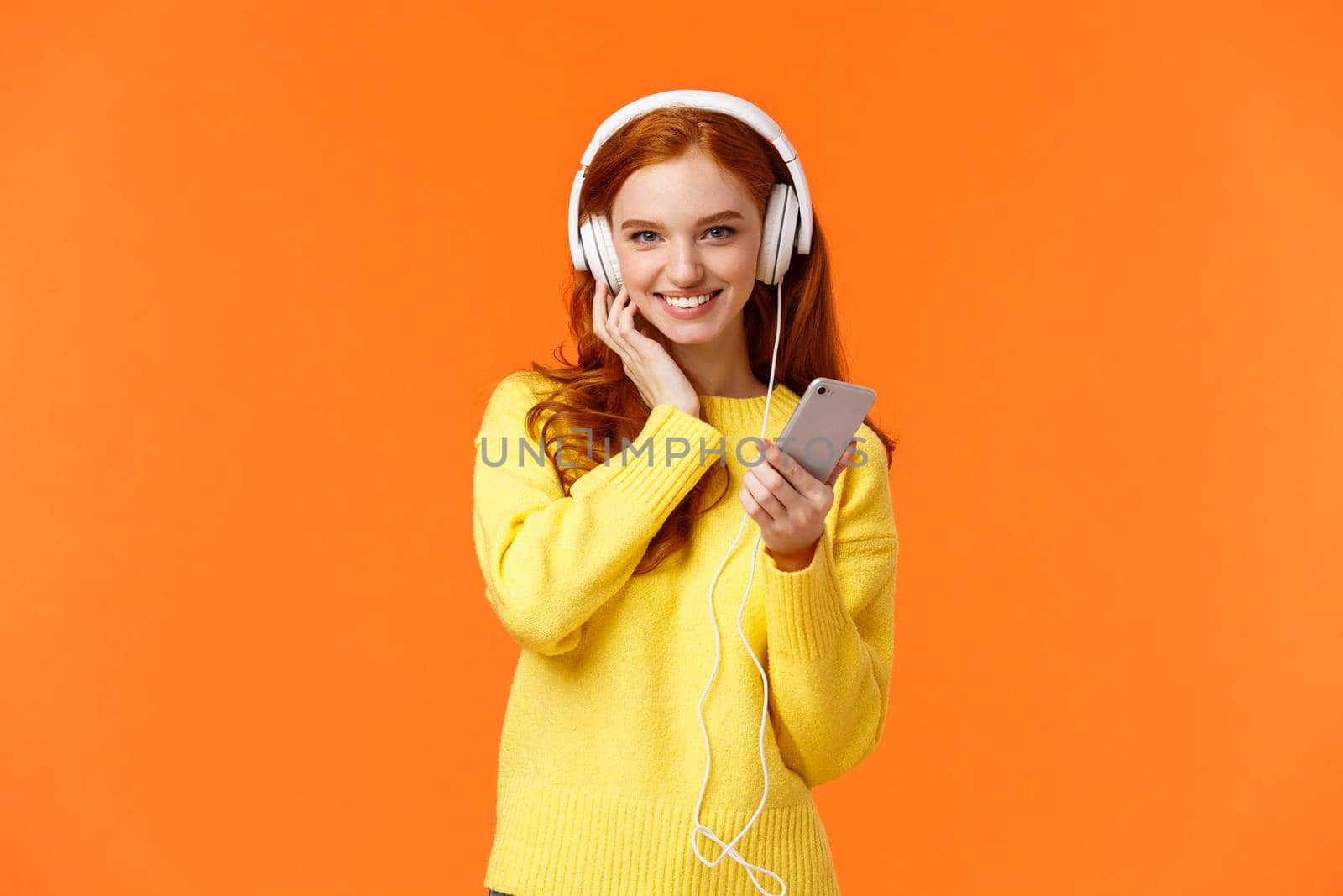 Sassy good-looking redhead female in yellow sweater, listen music white headphones, touch earphones and holding smartphone, listen music, enjoy good earbuds quality, smiling, orange background.