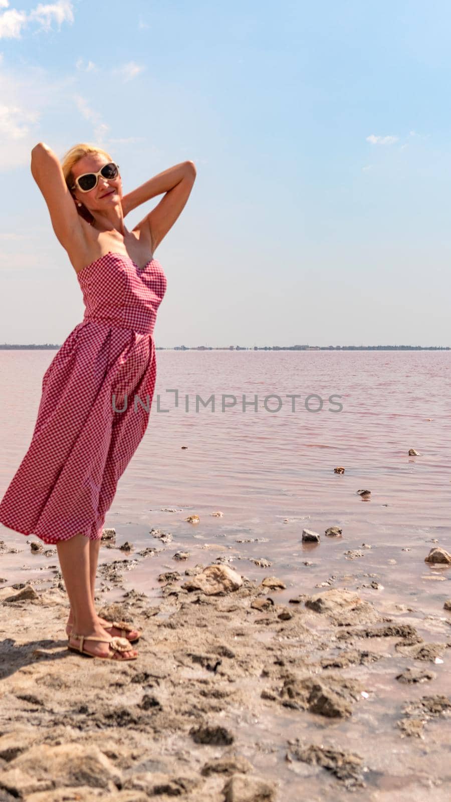 Cheerful lady in a red dress young smiling at the camera against the background of a red lake with blue clouds in hot sunny weather