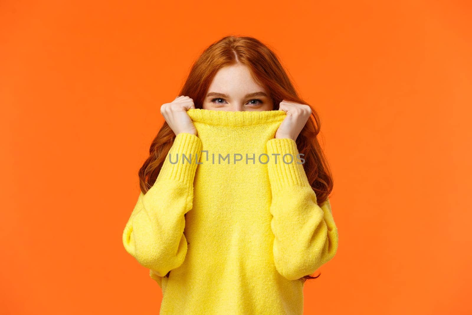 Girl hiding from winter cold in her warm soft sweater. Cute redhead woman pulling collar on nose and peeking at camera with smiling eyes, glancing at you happy, orange background by Benzoix