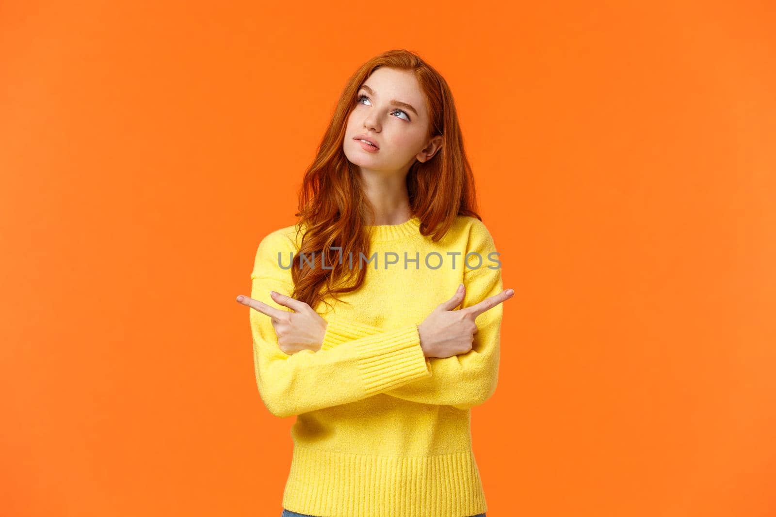 Pensive girl trying make decision, thinking, looking upper left corner thoughtful, pondering what buy for holidays, pointing sideways left and right, looking up dreamy, orange background.