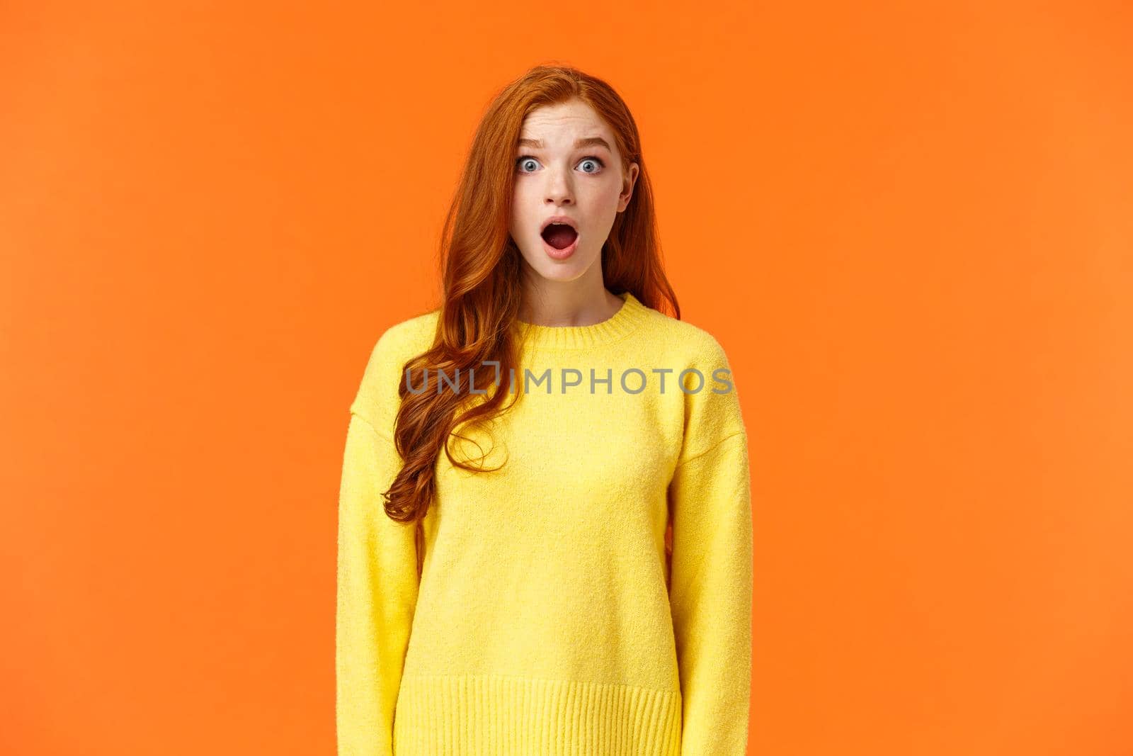 Shocked redhead cute girl drop jaw astounded and speechless, stare camera popped eyes, cant believe, standing impressed and fascinated, losing speech over orange background.