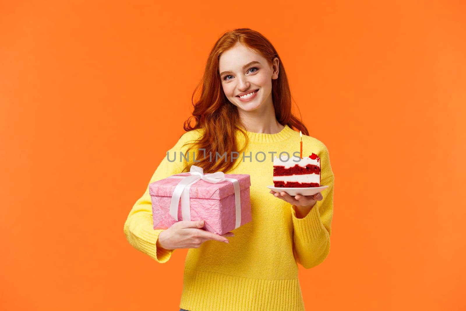 Cute redhead girl coming party with wrapped gift and piece of delicious b-day cake, asking friend blow-out candle and make with celebrating girlfriends birthday, smiling over orange background.