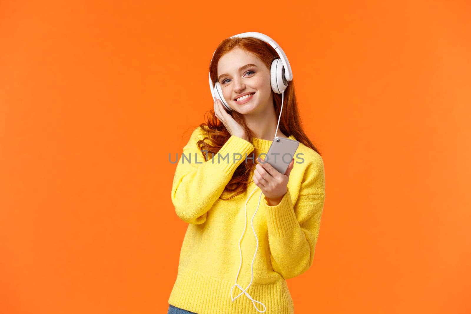 Waist-up portrait cheerful lovely young woman with red hair listen music in headphones, tilt head and smiling satisfied, holding smartphone, pick song for tender calm evening, orange background.