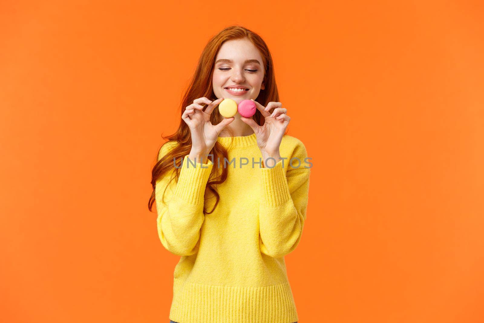 Redhead silly and happy gorgeous girl buy tasty macarons, holding food and looking with desire and affection, like sweets, want eat them, standing orange background satisfied and joyful by Benzoix