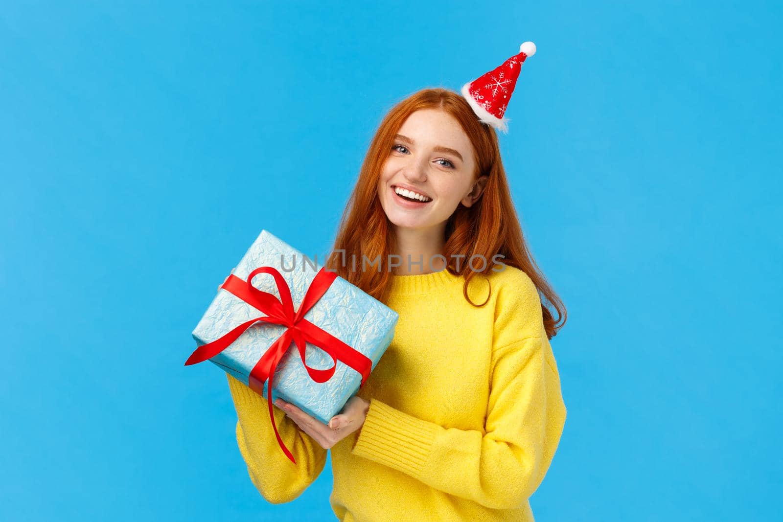 Holidays, celebration and presents concept. Cheerful redhead girl love christmas, enjoy celebrating with friends, receive gift in wrapped box from secret santa, wearing cute hat, blue background.