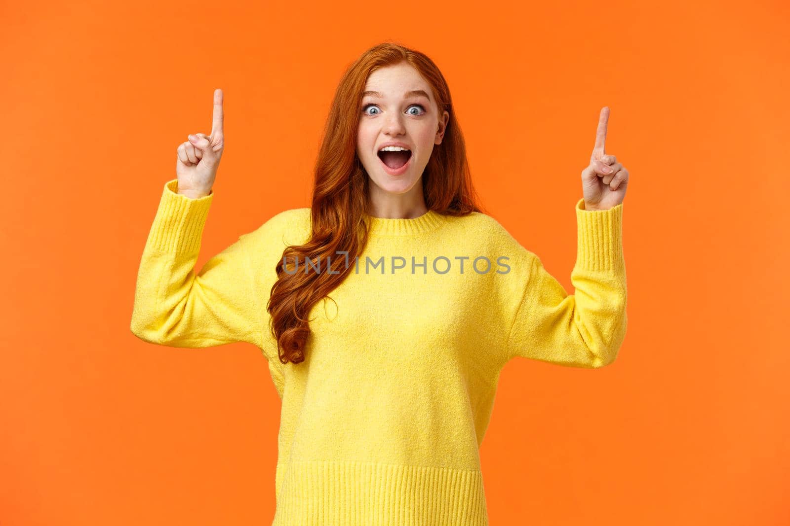 Excited girl travel abroad during winter holidays, time pack luggage. Astonished and cute redhead woman express happiness, pointing up at sale, promotion and smiling fascinated, orange background by Benzoix