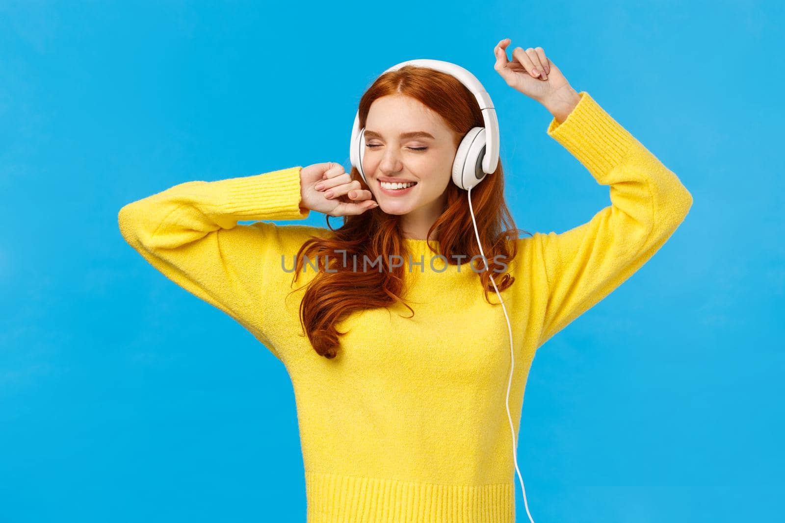 Carefree charming redhead female in yellow sweater, dancing relaxed and joyful with hands lifted up, close eyes and smiling happy, listen music headphones, standing blue background.