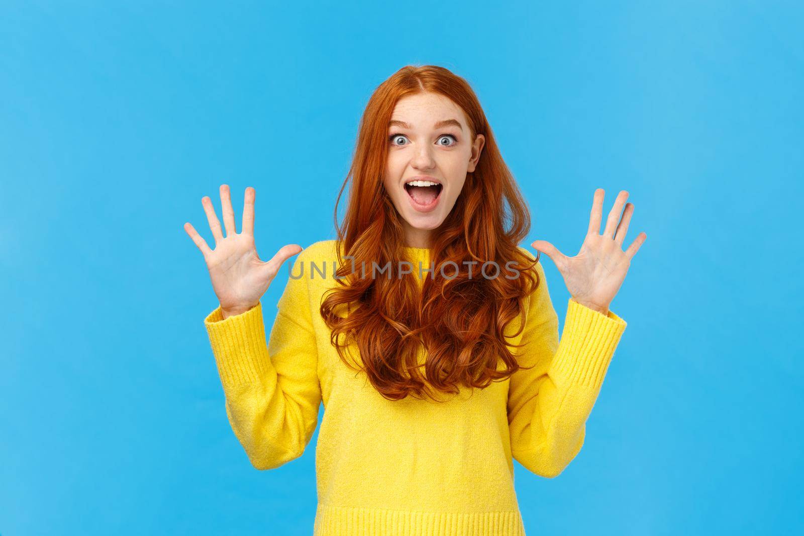 Excited, overwhelmed happy redhead cute girl in yellow sweater, screaming from joy telling about christmas gifts, showing ten fingers, order dozen products, standing joyful blue background.