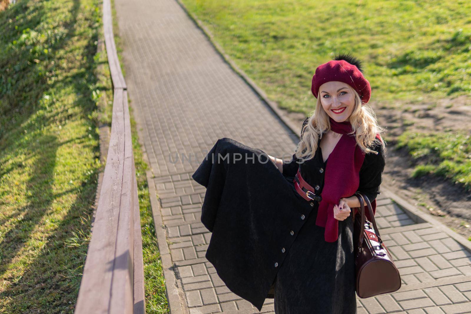 A joyful girl walks up the stairs in burgundy palette and biret, looks at the camera in black clothes, in the fall against the background of a pond there are blue clouds. by 89167702191