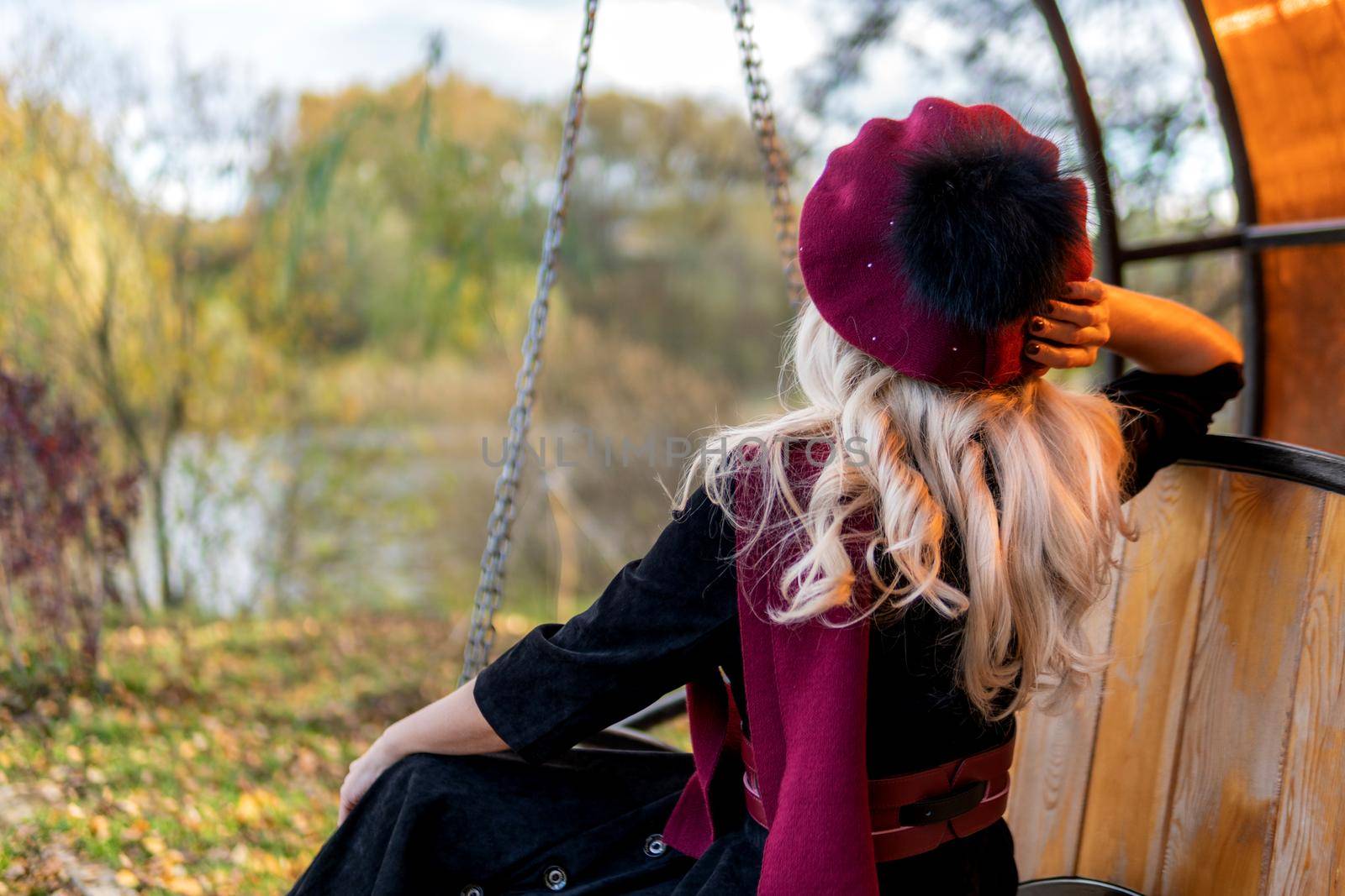 A woman sits with her back on a garden swing, tossed in a burgundy coat and in a biret, with beautiful eyes, in autumn against the background of the pond blue clouds. by 89167702191