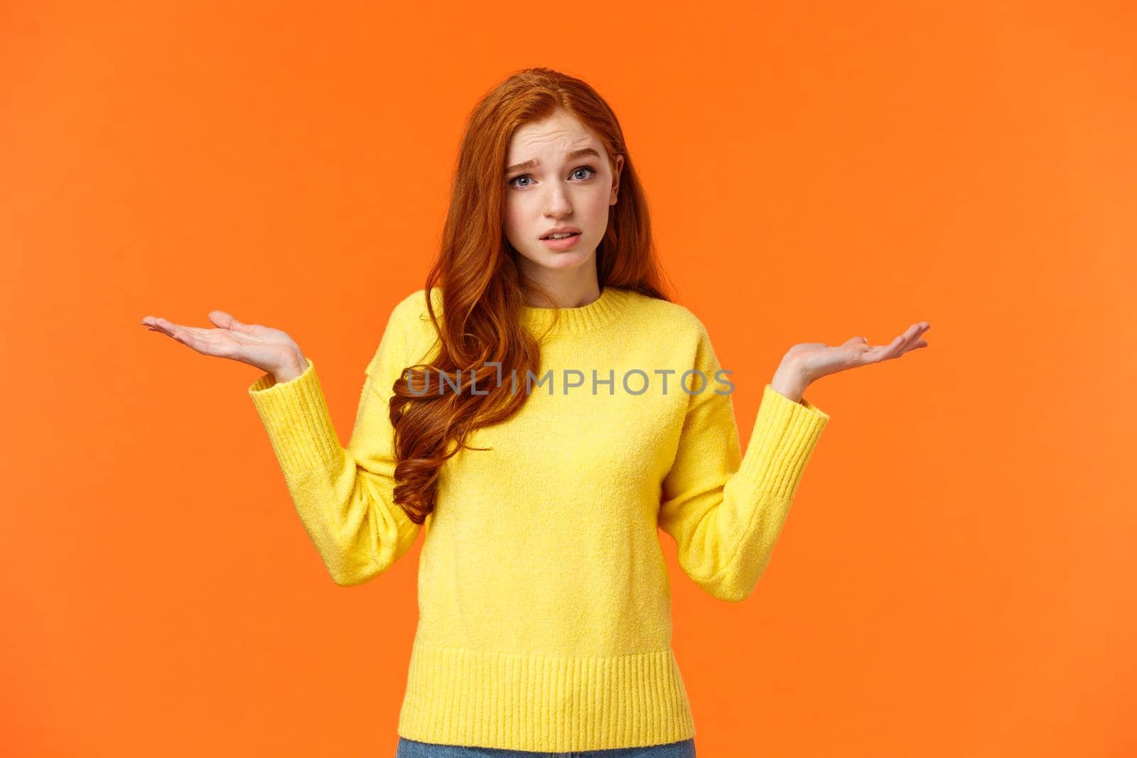 Perplexed cute redhead girl, shrugging with raised arms and looking confused, dont know answer, cant help, feeling confused or indecisive, cannot understand, standing orange background.