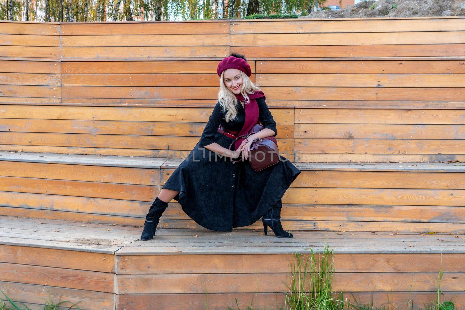 A joyful woman sits on a summer theater bench, made of wood in a burgundy coat and biret, in the fall against the blue sky. by 89167702191