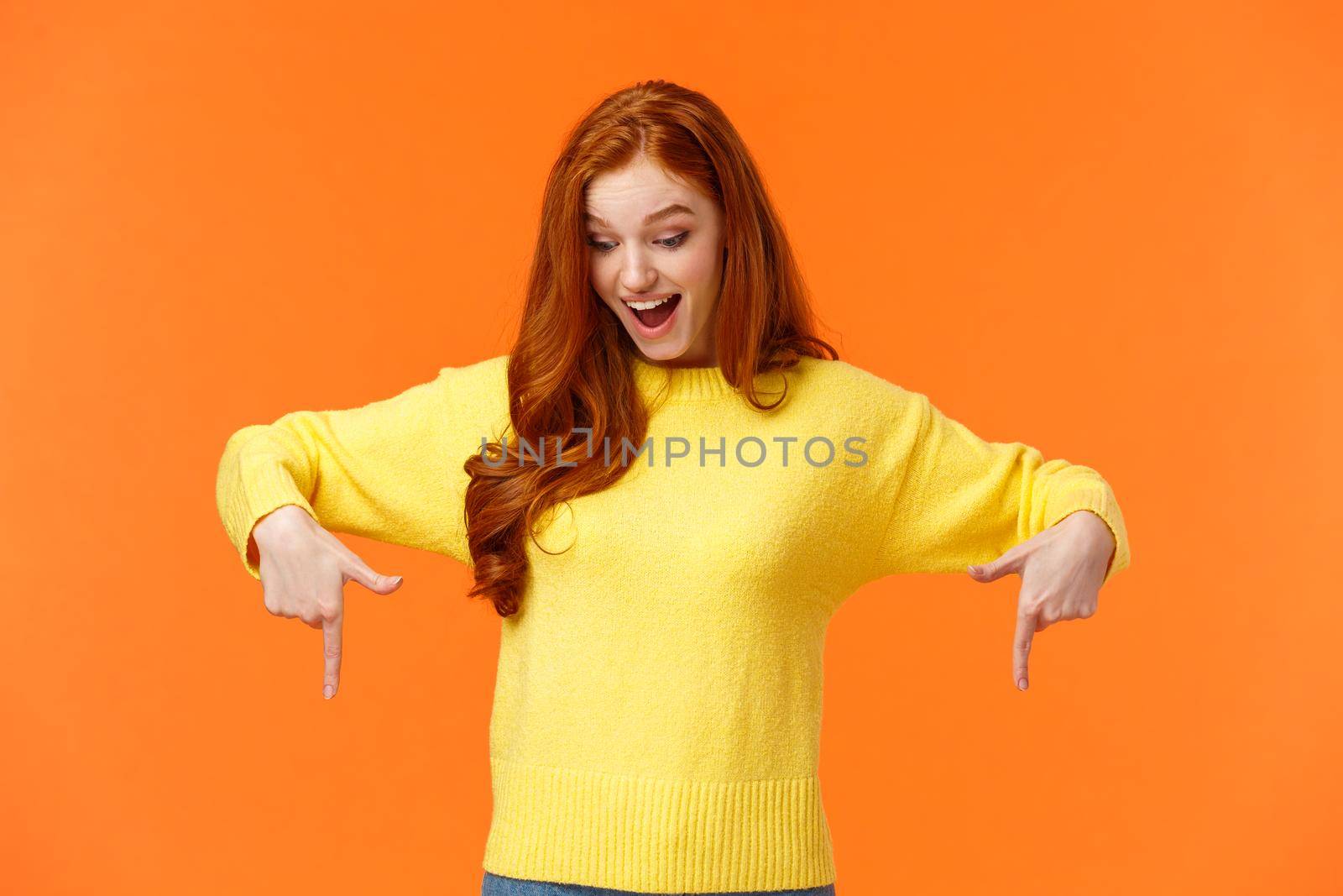 Wondered and excited, amused cute redhead woman in yellow sweater pointing fingers down, look bottom and smiling, check-out product promo, winter holiday event, orange background by Benzoix