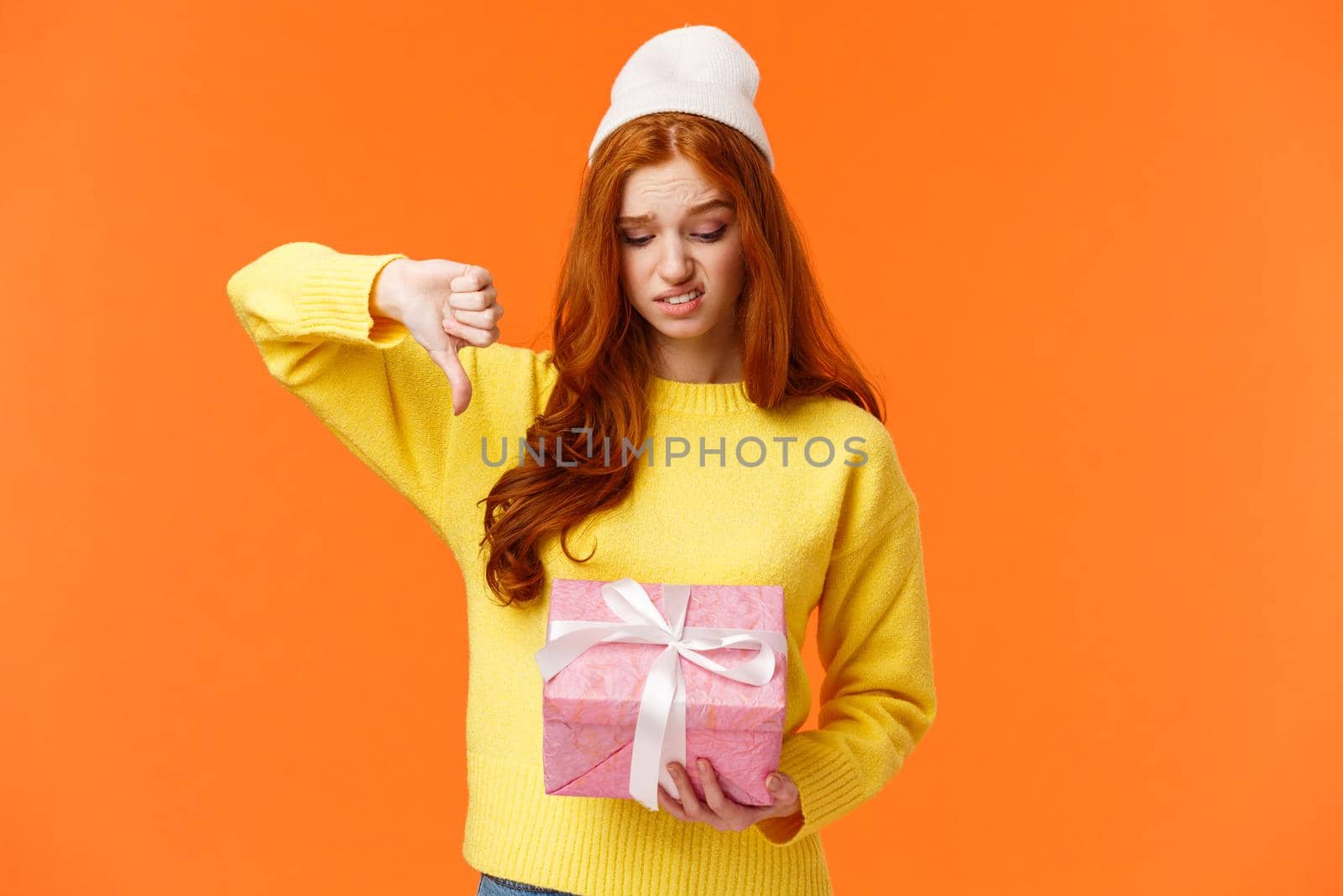 This gift sucks. Displeased problematic teenager, girl dont like present, looking at box with unsatisfied, displeased expression, grimacing skeptical, show thumb down in dislike, orange background.