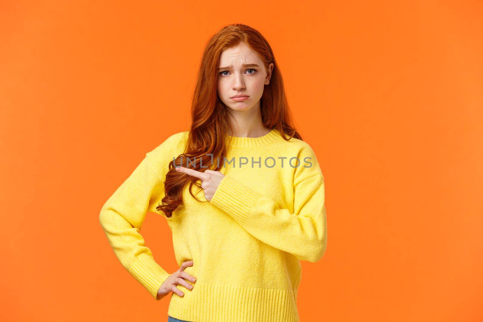 Girl looking upset and unsatisfied. Sad sulky redhead woman pointing left with envy or jealousy cant make it to party, frowning and sulking, showing what she want, standing orange background unhappy by Benzoix