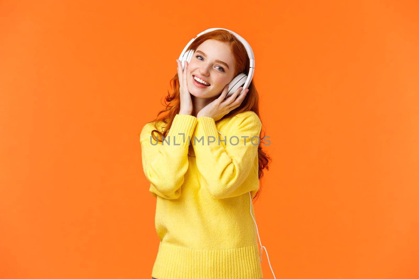 Waist-up portrait cute teenage redhead girl in white headphones, tilt head listen music, touch earphones and smiling camera, singing along favorite song, standing orange background. Copy space