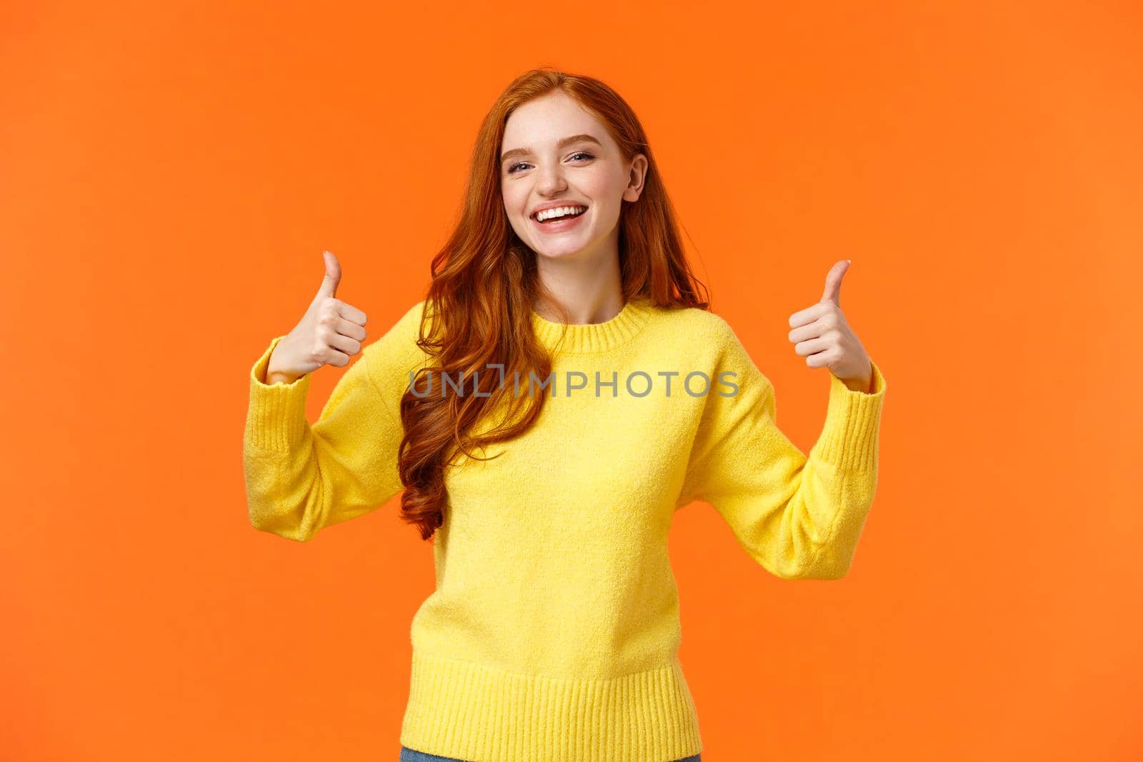 Holidays, gestures, people concept. Cheerful cute redhead girl smiling and showing thumbs-up recommend product, give positive reply, agree or like something, nod agreement, orange background.