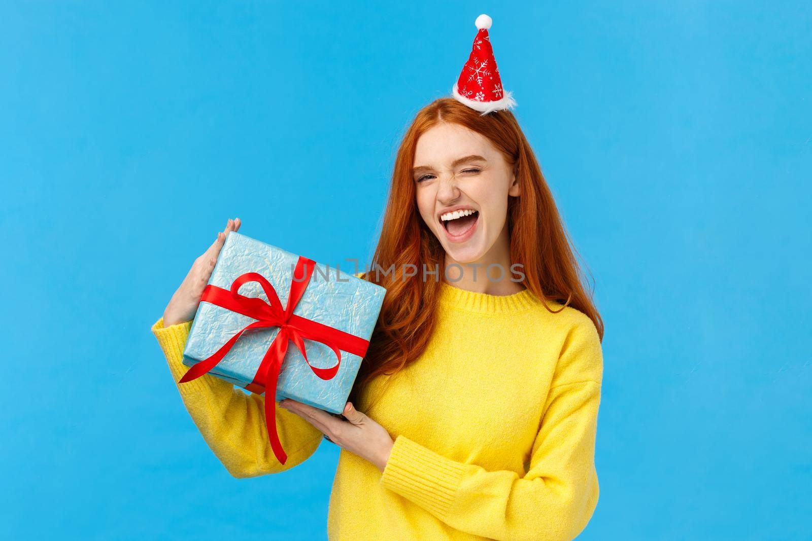Proud girl wrapped gift for girlfriend herself, winking sassy and pleased, smiling joyfully, wearing christmas hat, holding box with present and congratulating friend happy holidays, bllue background.