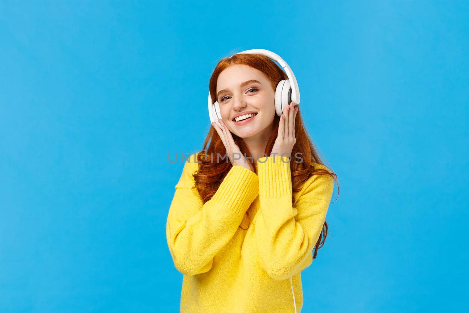 Fashionable attractive, carefree redhead woman in yellow sweater listening music with headphones, smiling tilting head joyfully, receive new earphones as christmas gift, standing blue background.