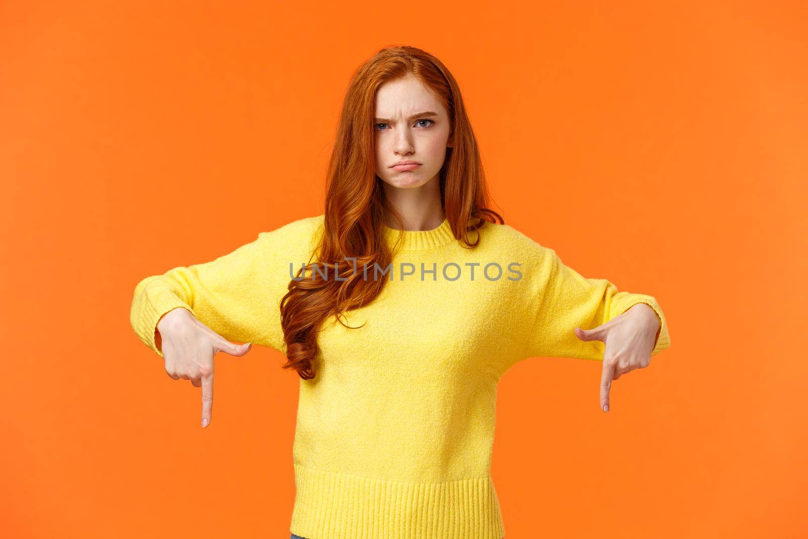 Mad unsatisfied cute redhead girlfriend, ginger girl in yellow sweater, frowning disappointed and angry, pointing down sulking, feeling offended with disapproval, standing orange background.