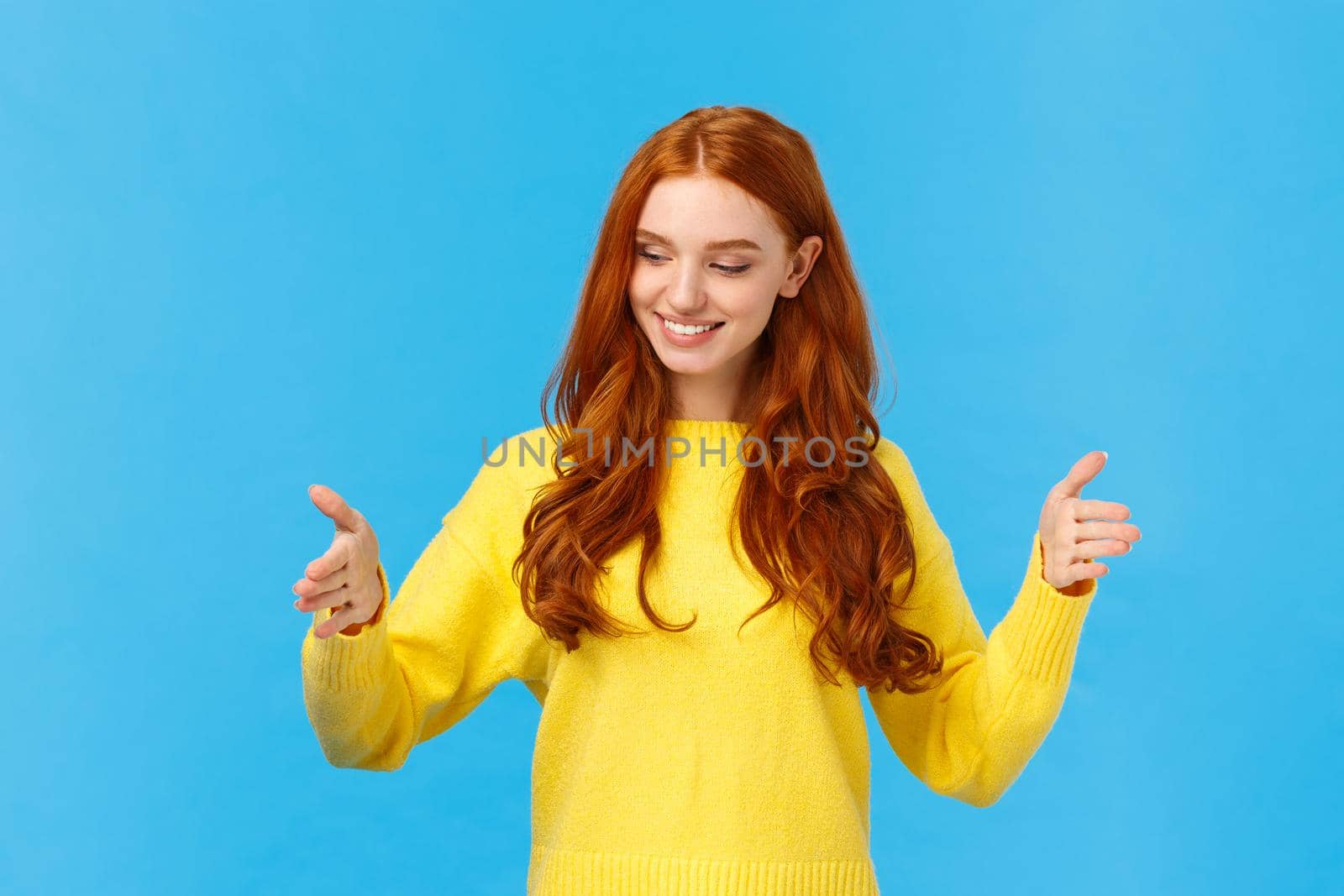 Happy good-looking young woman with red curly hair, freckles, shaping long object as if holding box over chest, looking satisfied, pleased with size, explain how big was gift, blue background.