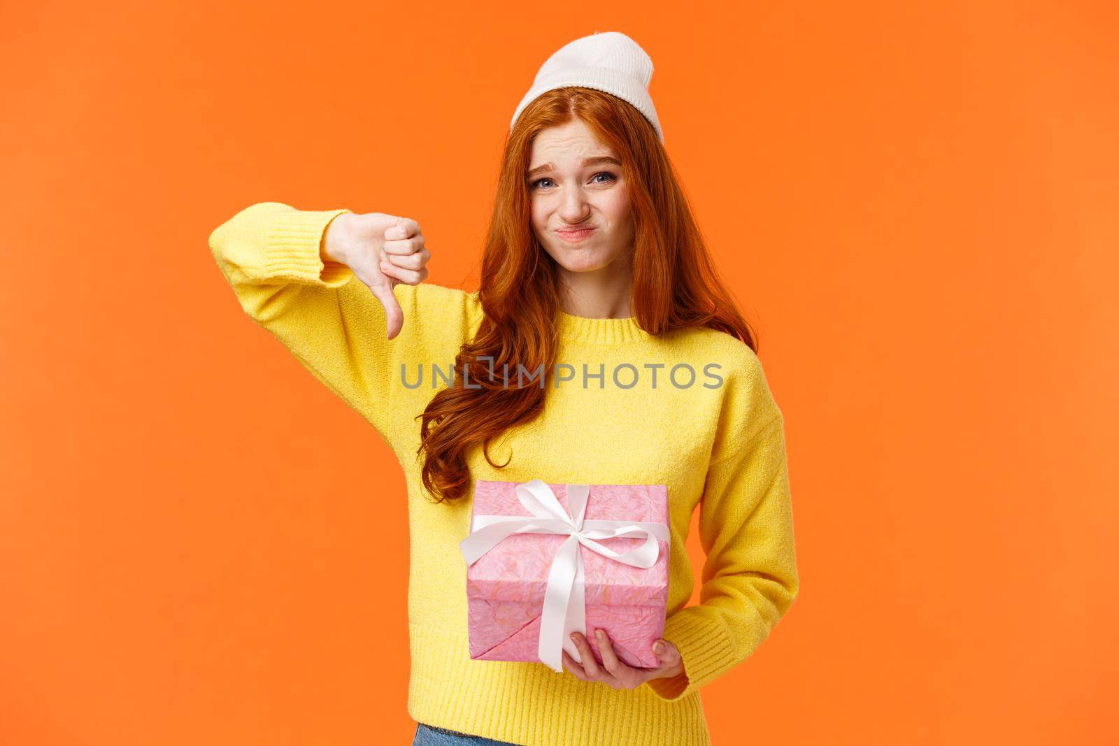 Skeptical and awkward cute redhead picky girl dont like christmas gift from aunt, show thumb-down, grimacing with uncomfortable judgemental expression, holding pink ugly present, orange background.