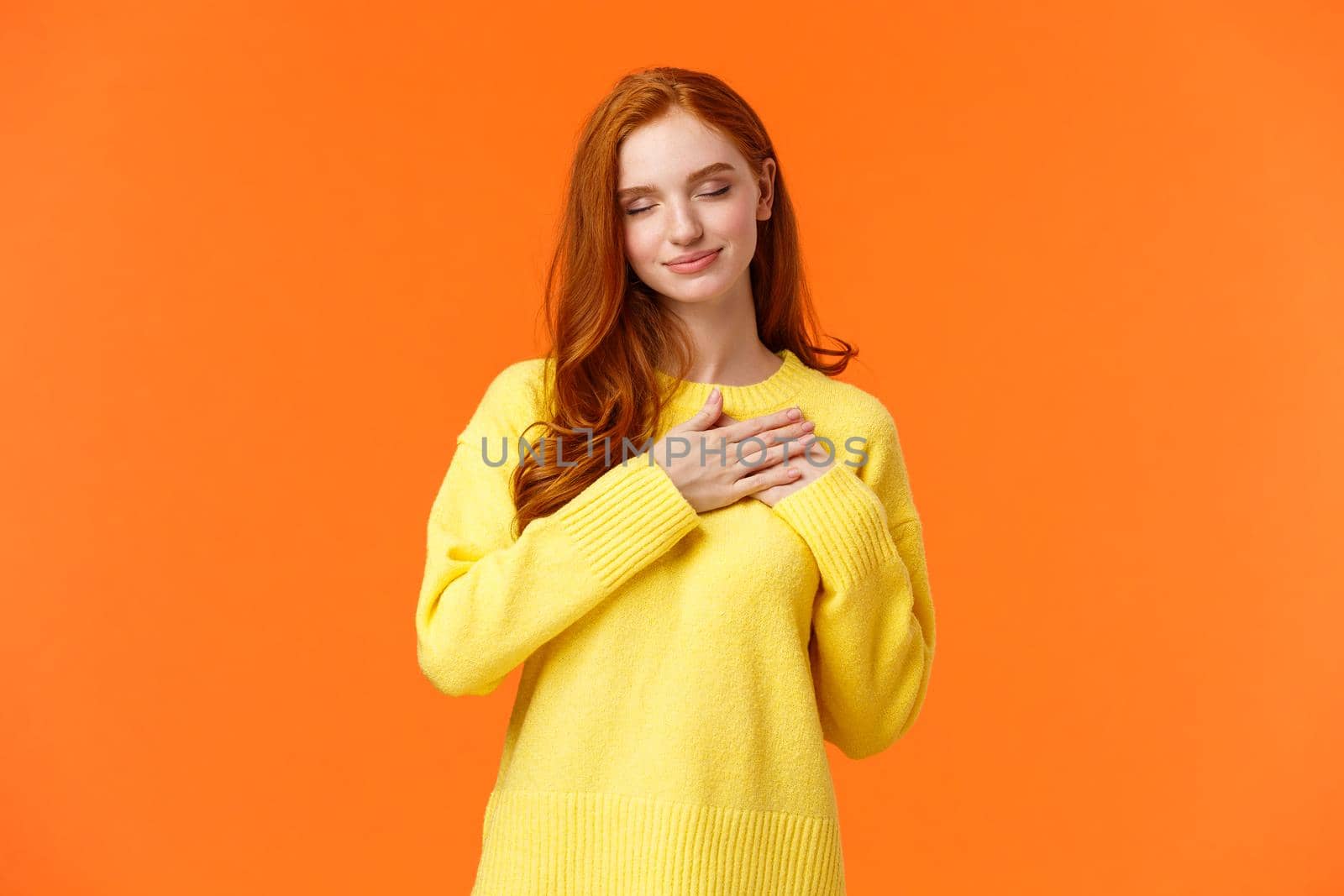 Tender and romantic, lovely young modern redhead woman 20s, touch heart and close eyes, remember dearest, touching moment, recall perfect date, keep memories in soul, orange background.