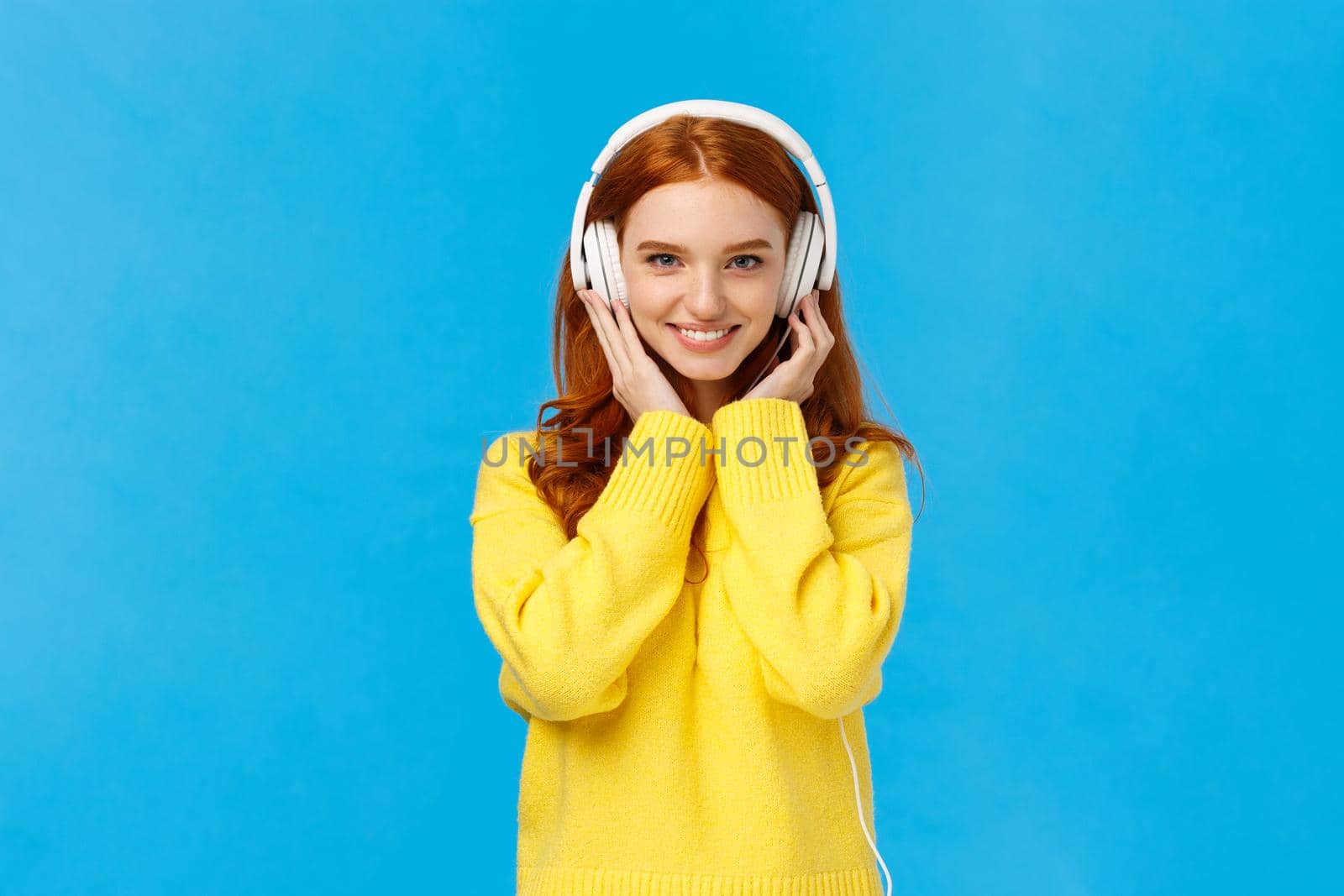 Waist-up shot sassy good-looking redhead, ginger girl with blue eyes and freckles in soft yellow sweater, listen music headphones, press earphones to ears, smiling hear new song favorite singer.