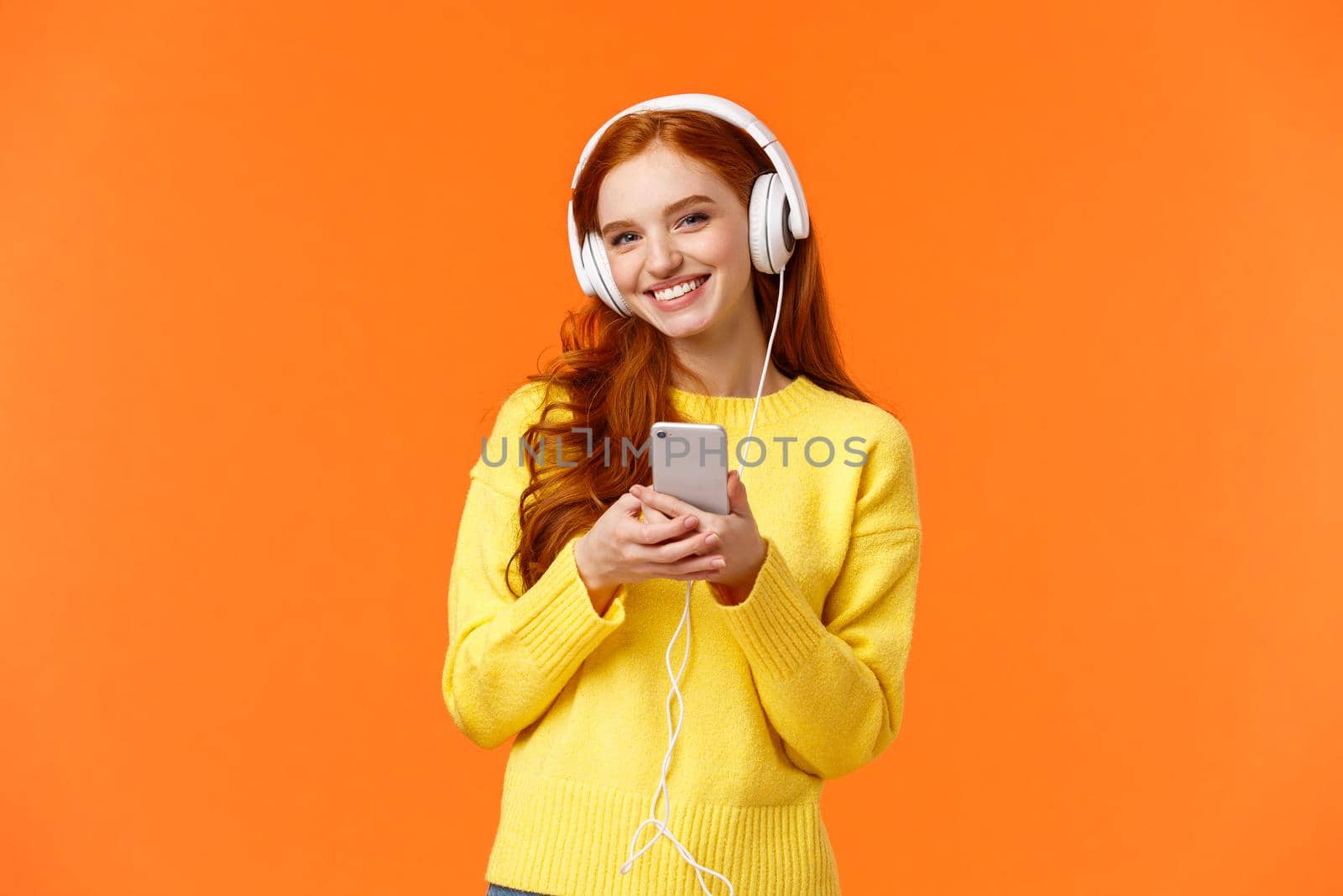 Technology, shopping and people concept. Modern cute hipster redhead girl wearing white headphones, holding smartphone, smiling carefree, enjoy nice music sound, standing orange background.