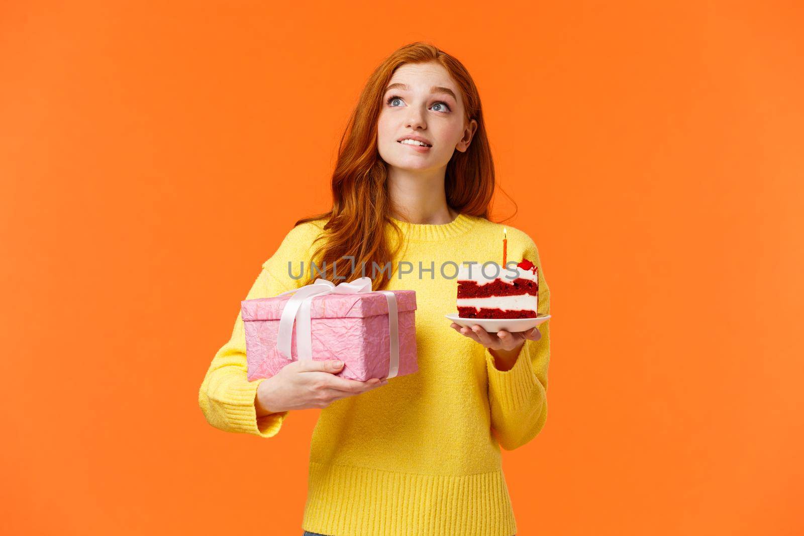 Dreamy, silly cute redhead girl with passionate, admiring look, biting lip have desire or wish, holding piece b-day cake delicious dessert, blowing candle, receive birthday gift in box, orange wall.